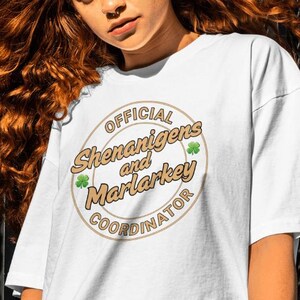 Könnte beinhalten: Weißes T-Shirt mit einem braunen Kreis-Motiv, auf dem "Official Shenanigans and Marlarkey Coordinator" mit grünen Kleeblättern steht.