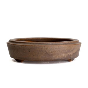 May include: A round, shallow, brown ceramic bonsai pot with a matte, textured finish. The container features a wide, flat rim and is supported by small, integrated feet at the base, presenting a simple, functional design for plant cultivation.