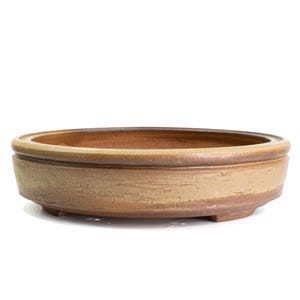 May include: A shallow, round ceramic bonsai pot with a textured, earthy tan and brown glaze. The vessel features a wide, flat rim and a slightly tapered body that rests on small, integrated feet, showcasing a natural, unrefined clay finish.