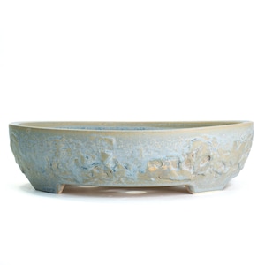 May include: A light blue oval-shaped ceramic planter with a textured surface. The planter has a raised, decorative pattern around the sides and sits on three small feet. The interior is a lighter shade of blue.