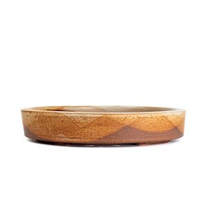 May include: A shallow, circular ceramic bonsai pot with a low profile and a flat base. The exterior features a variegated glaze with earthy tones of tan, beige, and dark brown, displaying a textured, mottled finish across the entire surface.