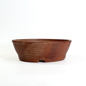 May include: A brown, oval-shaped bonsai pot with a textured, ridged surface. The pot has a small drainage hole on the side and is made of ceramic. The pot is designed for planting small trees and plants.