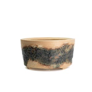 May include: A beige ceramic planter with a textured, dark green and brown band around the middle. The planter has a rounded oval shape and sits on three small feet. The interior is a lighter beige color.