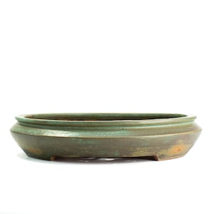 May include: A shallow, oval-shaped ceramic bonsai pot with a matte, weathered finish in shades of muted green and earthy brown. The vessel features a wide, flat rim and a tapered base with small, integrated feet, showcasing a textured surface.