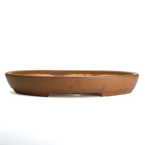 May include: A brown, oval-shaped ceramic bonsai pot. The pot has a low profile with a slightly textured surface and a rectangular foot on each end. The interior is a lighter shade of brown. The pot is designed for bonsai trees.