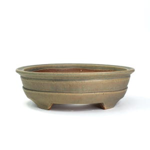 May include: An oval-shaped bonsai pot in a muted green-brown hue. The ceramic pot features a series of horizontal ridges around its body and rests on three small feet. The interior has a reddish-brown color.