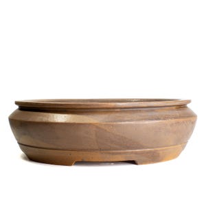 May include: A shallow, oval-shaped ceramic bonsai pot with a smooth, matte finish in earthy shades of brown and tan. The vessel features a wide, flat rim and a slightly tapered base with small cutouts along the bottom edge for drainage.