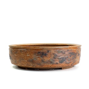 May include: A round, shallow ceramic bonsai pot with a textured, earthy brown surface featuring mottled dark grey and blue glaze accents. The vessel has a raised rim, a flat base with small feet, and a wide, open top for planting purposes.