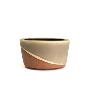 May include: A small, handcrafted ceramic bowl with a unique color scheme. The bowl features a diagonal design with a warm brown base, transitioning to a muted gray-green, and a light green rim. The interior is dark.