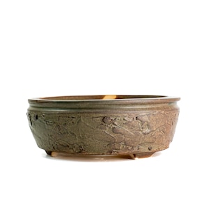 May include: A round, shallow ceramic bonsai pot with a textured, earthy olive-green exterior finish. The vessel features a smooth, slightly raised rim and rests on small, integrated feet, showcasing a rustic, hand-crafted appearance.