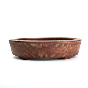 May include: A brown, oval-shaped bonsai pot with a textured, vertical-line pattern. The ceramic pot has a low profile and sits on three small feet. The pot is designed for planting small trees and plants.