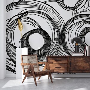 Può includere: Un interno di un soggiorno moderno con una parete bianca con un design di carta da parati astratto in bianco e nero di cerchi sovrapposti. Una sedia in legno con una seduta in vimini intrecciato si trova di fronte a una credenza in legno scuro con due cassetti. Una lampada nera con un paralume nero si trova sopra la credenza.