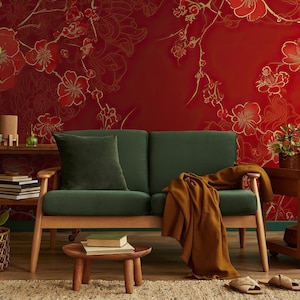 Può includere: Un divano in velluto verde con braccioli e gambe in legno si trova di fronte a un muro rosso con un motivo floreale dorato. Il divano ha una coperta di lana marrone drappeggiata sopra di esso. Ci sono due piccole piante in vaso su un tavolo di legno accanto al divano.