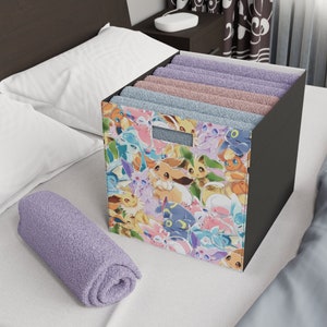 Puede incluir: Un cubo de almacenamiento negro con un estampado colorido de personajes de Pokemon de dibujos animados, incluyendo Eevee, Espeon y Umbreon. El cubo está lleno de toallas dobladas en tonos de morado, rosa y azul.