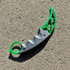 May include: A gray and green 3D printed karambit knife with a textured handle and a green ring.