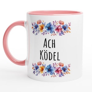 Könnte beinhalten: Weiße Keramiktasse mit rosafarbenem Rand und Henkel. Die Tasse zeigt ein Blumenmuster mit blauen, rosa und gelben Blumen. Der Text "ACH KÖDEL" ist in Schwarz auf die Tasse gedruckt.