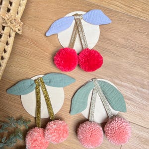 Könnte beinhalten: Drei dekorative Kirschornamente. Jede Kirsche hat ein Paar flauschiger Pomponkugeln in Rot und Rosa, die mit glitzernden goldenen oder silbernen Stielen und Stoffblättern in Blau und Grün an einem Holzring befestigt sind.