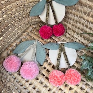 Könnte beinhalten: Dekorative Kirschornamente mit rosa Pompons, grünen Stoffblättern und goldglitzernden Stielen. Die Kirschen sind an einem weißen Holzring befestigt. Die Ornamente sind auf einem geflochtenen Korb angeordnet.
