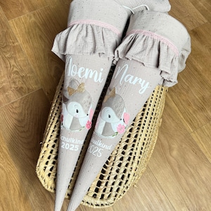 Könnte beinhalten: Zwei beige Schultüten mit einem Fuchsmotiv und den Namen "Moemi" und "Mary" aufgedruckt. Die Tüten haben einen gerüschten oberen Rand mit rosa Besatz und sind mit einer Fuchsgrafik und dem Text "Schulkinder 2025" verziert. Sie liegen in einem Weidenkorb.
