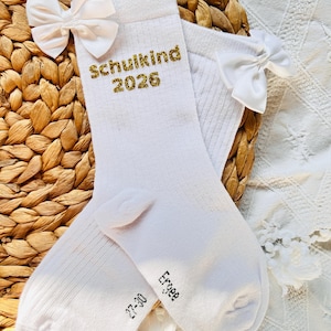 Könnte beinhalten: Ein Paar weiße Socken mit goldglitzerndem Text "Schulkind 2026". Jede Socke hat eine weiße Schleife. Die Socken haben die Größe 27-30. Sie liegen auf einem gewebten Hintergrund.