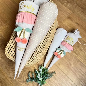 May include: Several fabric school cones in a woven basket. The cones are in various colors and patterns, including white with gold stars, and white with pink ruffles. One cone has the name "Emily" in gold script, and another has "Lilly". Each cone has a cherry decoration.