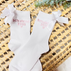 Könnte beinhalten: Ein Paar weiße Socken mit einer Satinschleife an der Außenseite jedes Bündchens. Die Socken sind mit dem rosafarbenen Schriftzug "Schulkind 2026" bedruckt, gefolgt von einem kleinen rosafarbenen Herzsymbol, das mittig unter dem Text platziert ist.