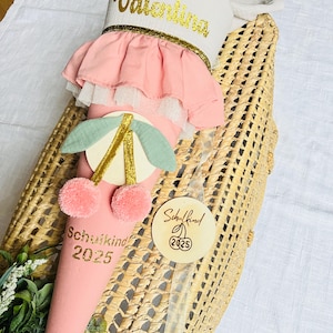 Peut inclure: Un cornet d'école rose avec le nom "Valentina" en lettres dorées. Le cornet présente un volant rose, un motif de cerise et le texte "Schulkind 2025". Une étiquette en bois avec une cerise et l'année 2025 est attachée.