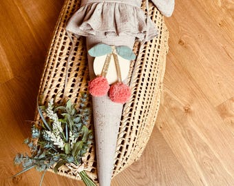 Pode incluir: Um cone de tecido cinza e branco com uma parte superior franjada e o nome "Livia" bordado em glitter dourado. O cone é decorado com um desenho de cereja e dois pompons cor-de-rosa. O cone está sentado numa cesta tecida.