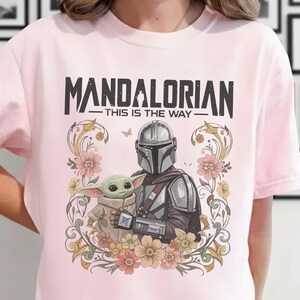 May include: Light pink t-shirt featuring a graphic of The Mandalorian and Grogu surrounded by floral elements. The text "MANDALORIAN - THIS IS THE WAY" is printed above the characters. The shirt has short sleeves.