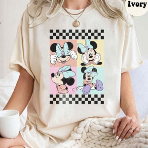 May include: A white t-shirt featuring a checkered pattern and a collage of four images of Minnie Mouse in various poses. The images are in a pastel colour palette and have a vintage look. The t-shirt is labelled "Ivory".