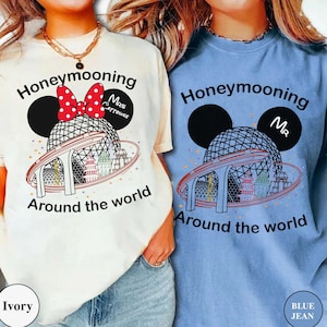 Op de afbeelding: Twee witte T-shirts met een Mickey Mouse-orenontwerp en de tekst "Honeymooning Around the World". Eén shirt zegt "Mrs. Mouse" en het andere zegt "Mr.". De shirts zijn op modellen die jeans dragen. Het shirt links is ivoor en het shirt rechts is blauw.