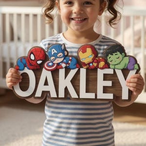Könnte beinhalten: Ein Namensschild aus Holz mit dem Namen OAKLEY in großen weißen Buchstaben, verziert mit bunten Cartoon-Illustrationen von Spider-Man, Captain America, Iron Man und Hulk in schlafender Pose, gehalten von einer Person in einem gestreiften Shirt.