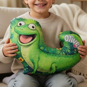 May include: A green dinosaur-shaped pillow with a smiling face and the name "Oliver" printed on it. The pillow has a tropical floral pattern and is being held by a smiling child. Wooden toy blocks are in the foreground.