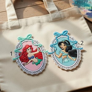 May include: A cream-colored tote bag featuring two embroidered patches. One patch depicts a red-haired mermaid in a pink oval frame, and the other shows a woman with black hair in a blue oval frame. The bag has two handles.