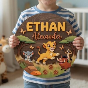 May include: A circular wooden wall sign featuring a layered design with the name Ethan Alexander in yellow and brown text. The artwork depicts a cartoon lion cub, a warthog, and a meerkat in a savanna setting with trees, grass, and small butterflies.