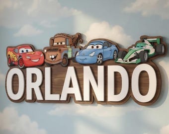 Może przedstawiać: Drewniana dekoracja ścienna z napisem "ORLANDO" w białych literach, z kolorowymi postaciami z kreskówek samochodów powyżej. Znak jest zamontowany na podstawie z ciemnego drewna, na tle błękitnej ściany z wzorami chmur. Białe łóżeczko dziecięce znajduje się na pierwszym planie.
