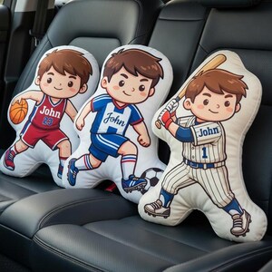 May include: Three cartoon-style pillows shaped like children playing sports. One pillow depicts a basketball player with the name "John" and the number 23. Another shows a soccer player with the name "John". The third pillow is a baseball player with the name "John" and the number 1.