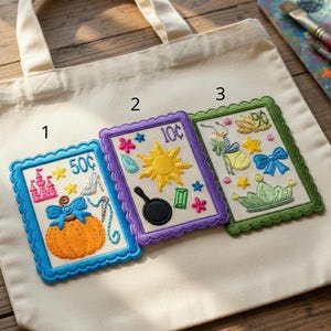 May include: Cream-colored tote bag with three embroidered patches. The patches feature fairytale-themed designs with a pumpkin, sun, and crown. The patches are framed in blue, purple, and green with decorative borders. A wooden palette and paintbrushes are in the background.