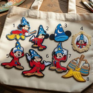 May include: A beige tote bag displays eight colorful embroidered patches. The patches feature a cartoon mouse in wizard attire, a blue wizard hat, and a framed portrait of the mouse. The patches are on a wooden surface with art supplies.