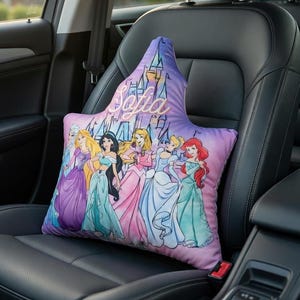 Op de afbeelding: Een decoratief autostoelkussen met Disney-prinsessen. Het kussen heeft een paarse en roze achtergrond met het woord "Sofia" in cursief. De prinsessen zijn afgebeeld in hun iconische jurken. Het kussen ligt op een zwarte autostoel.