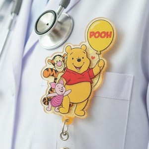 Op de afbeelding: Een gele en rode Winnie de Poeh thema badge reel. De badge toont Winnie de Poeh, Knorretje en Teigetje. Winnie de Poeh houdt een gele ballon vast met het woord "POOH" erop. De badge is bevestigd aan een witte jas met een stethoscoop.