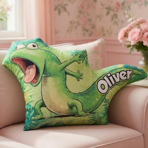 May include: A green cartoon lizard-shaped pillow with the name "Oliver" in white script. The pillow is on a pink couch in a room with floral wallpaper and a window. The lizard has its mouth open and tongue out.