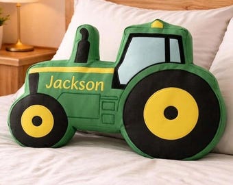 May include: A green tractor-shaped throw cushion featuring large black and yellow wheels, a light blue window section, and a black exhaust pipe. The side of the tractor is embroidered with the name Jackson in yellow lettering against the green fabric.
