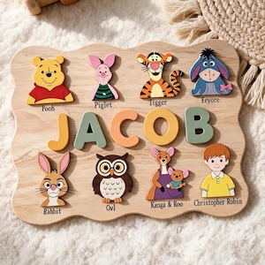 Op de afbeelding: Een houten puzzelbord met de naam JACOB in grote, gekleurde letters. Het bord bevat uitsparingen en illustraties van Winnie de Poeh-personages: Poeh, Knorretje, Teigetje, Iejoor, Konijn, Uil, Kanga en Roe, en Janneman Robinson.
