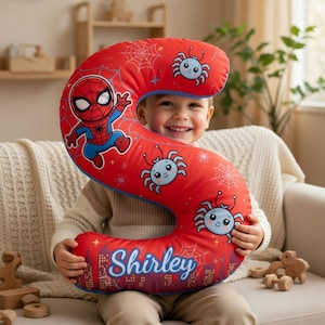 May include: A red, letter "S" shaped cushion featuring Spider-Man and cartoon spiders. The cushion has a blue border and the name "Shirley" is printed on the bottom. The background includes spiderwebs and a cityscape.