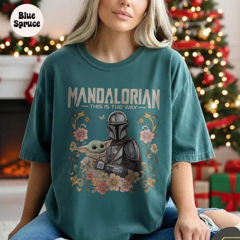 May include: Teal-colored t-shirt featuring the text "MANDALORIAN" above an illustration of the Mandalorian and Grogu, surrounded by floral accents. The phrase "THIS IS THE WAY" is below the text. The Blue Spruce logo is in the upper left corner.
