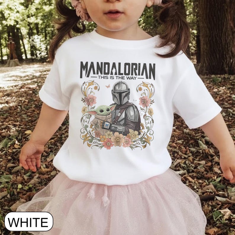 May include: White t-shirt featuring the text "MANDALORIAN - THIS IS THE WAY" above an illustration of the Mandalorian and Grogu surrounded by floral elements. The shirt is worn by a child.