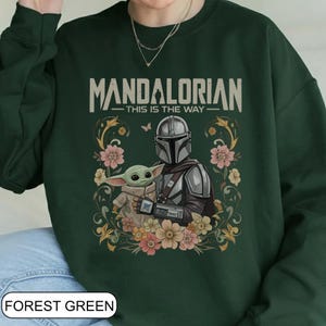 May include: Forest green sweatshirt featuring the text "MANDALORIAN - THIS IS THE WAY" above an illustration of the Mandalorian and Grogu surrounded by flowers. The sweatshirt is a solid forest green color.