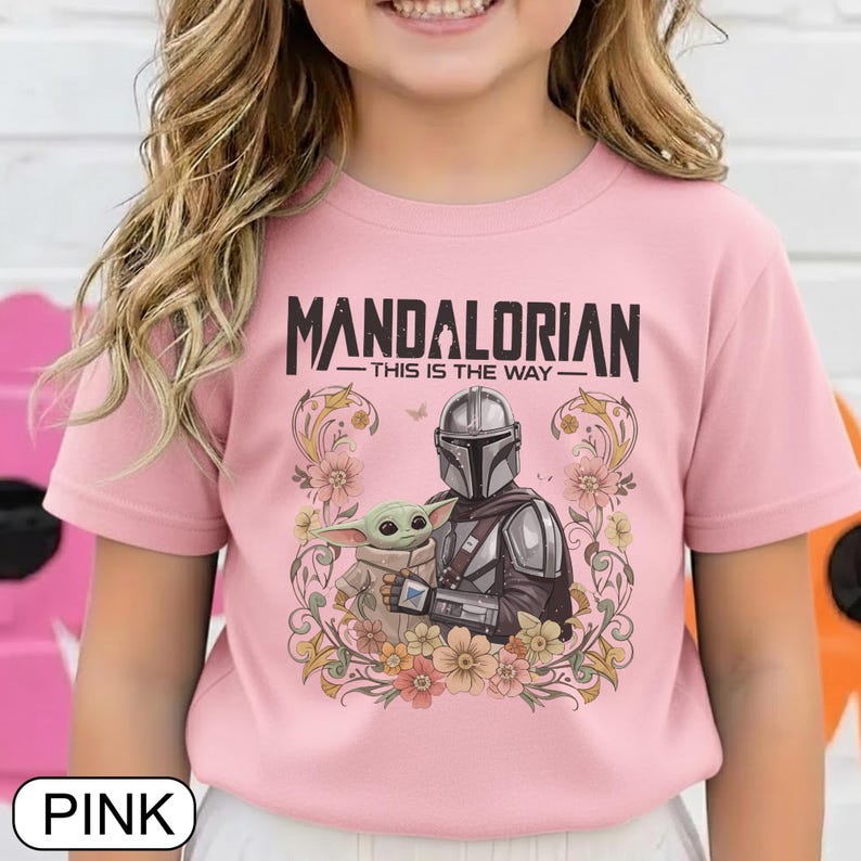 May include: Pink t-shirt featuring a graphic of The Mandalorian and Grogu, surrounded by floral elements. The text "MANDALORIAN - THIS IS THE WAY" is printed across the top. The shirt is a solid pink color.