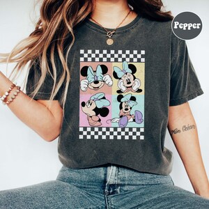 May include: A dark grey t-shirt with a chequered pattern and a graphic of four different images of Minnie Mouse. The graphic is in a retro style with pastel colours.